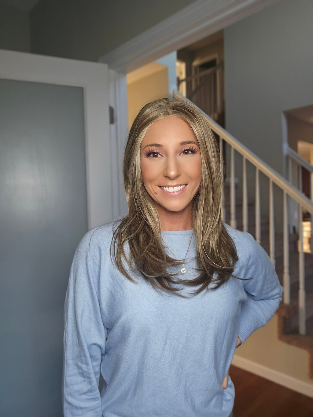 A smiling woman with blonde hair wearing a light blue long-sleeve top, standing in a home hallway with a staircase in the background.