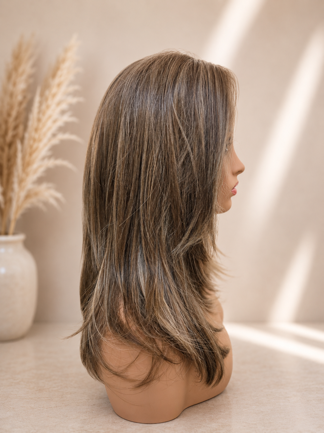 A shoulder-length layered wig with ash brown and light blonde highlights, displayed on a mannequin head in profile against a warm beige background with soft window light shadows.