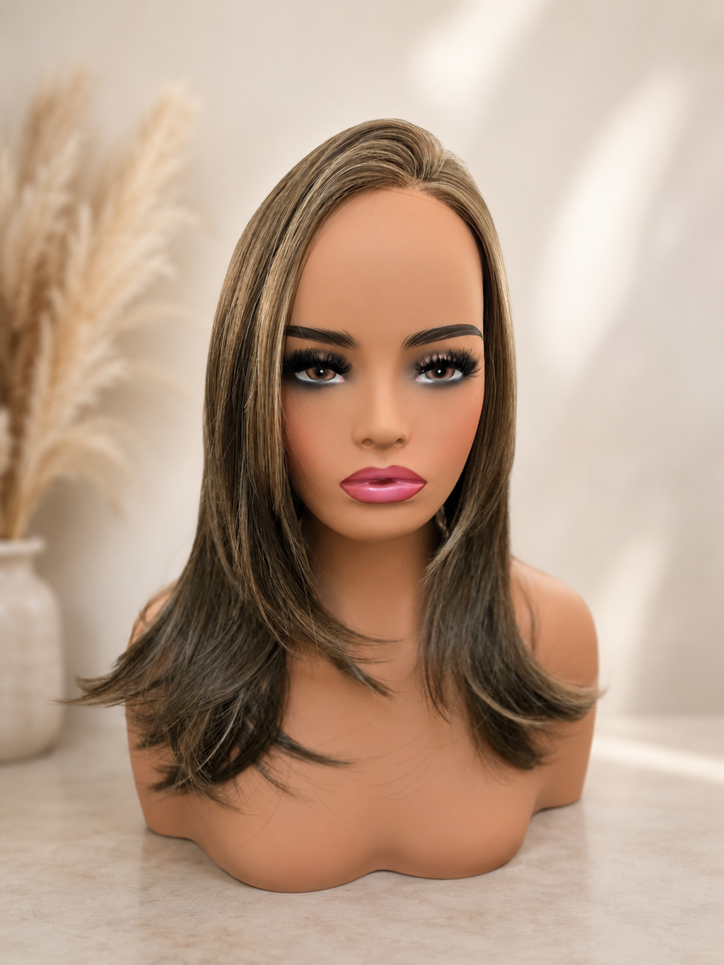 A shoulder-length wig with highlighted brown and blonde streaks, displayed on a mannequin bust against a softly lit neutral background with dried grass decor.