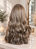 Long, wavy hair extensions or a wig shown from the back, featuring a blend of medium brown and warm blonde highlights, styled on a marble counter in a softly lit interior setting.