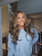 A smiling woman with long, wavy brown hair with blonde highlights, wearing a light blue knit sweater, standing in a home interior with a staircase in the background.